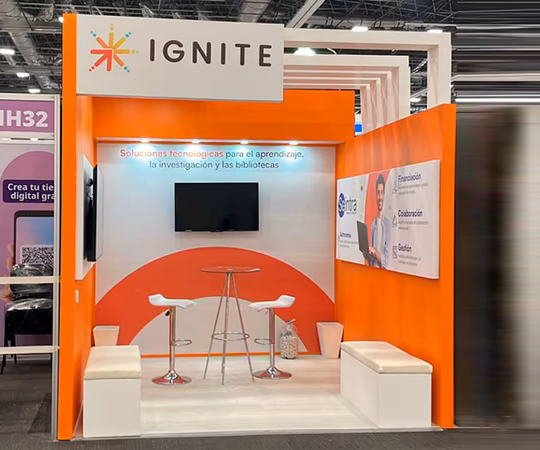 展区展示“IGNITE”品牌，两把白色椅子，一张小圆桌，一台挂在墙上的电视，以及橙色和白色的装饰，配有西班牙语的介绍性横幅。